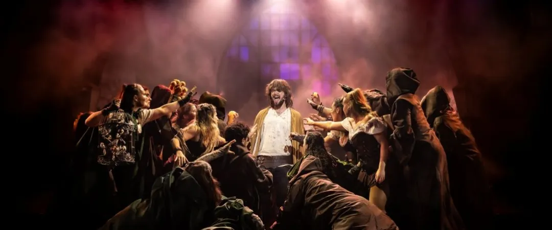 Dramatic stage scene with a man in the centre, surrounded by animated performers in costumes, under colourful spotlights, creating a tense atmosphere.