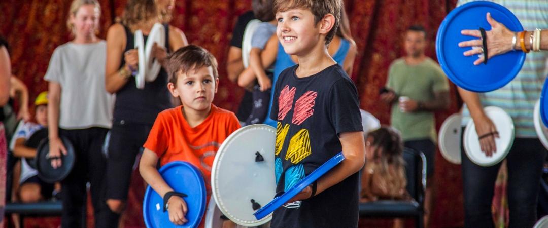 Children enthusiastically participate in a group activity, holding colorful blue and white discs. They stand in a room with a red curtain, smiling and engaged.
