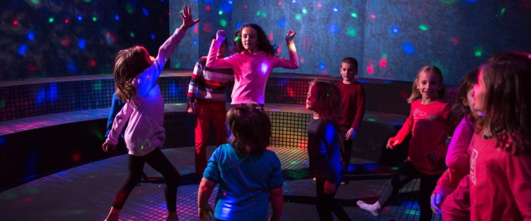 Children joyfully dance under colorful disco lights in a dark room with a curved bench. The scene is lively, capturing a playful and energetic atmosphere.