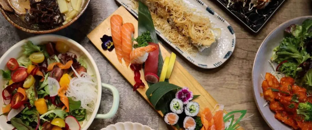 Overhead spread of Japanese cuisine: ramen, sushi, sashimi, colorful salad, takoyaki, and grilled dishes create an inviting feast.