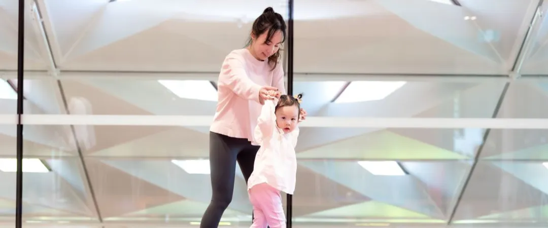 A woman assists a toddler walking on a balance beam in a bright gym. Both wear pink. 
