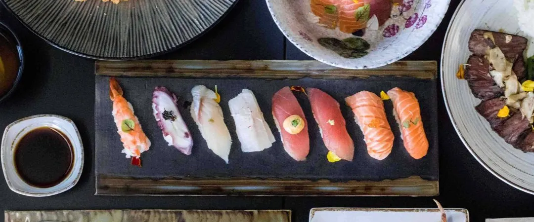 Overhead view of an elegant sushi spread: colorful nigiri, maki rolls, dumplings, tacos, dipping sauce, and salad on dark plates.