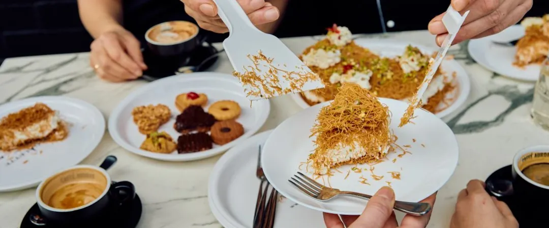 Hands serve a shredded pastry dessert onto a plate at a restaurant table, surrounded by coffee, pastries, plates, and diners in a warm communal setting.