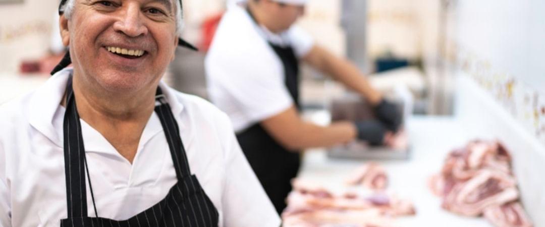 Smiling butcher in a black apron and hat stands in front of a meat counter, with a coworker cutting meat in the background, conveying a friendly atmosphere.