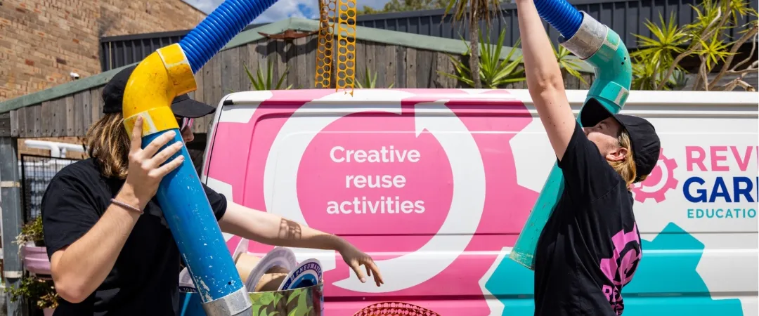 Two women in black tshirts and black hats hold colourful pipe in front of pink and blue van.