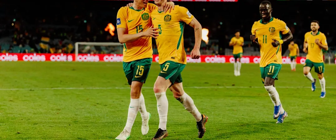 Two soccer players in yellow and green jerseys celebrate joyfully on the field, arms around each other, with teammates running behind them.