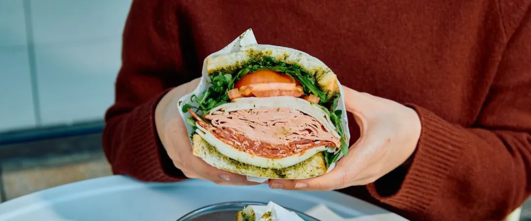 Person in a rust sweater holds a large deli sandwich layered with greens, tomato, and sliced meat over a plate beside coffee.