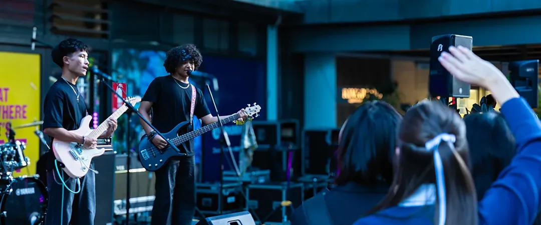 A band performs energetically on an outdoor stage, with a guitarist and bassist. The crowd is engaged, with a person raising their hand in excitement.