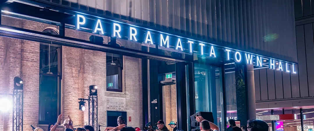 A lively crowd gathers under the illuminated "Parramatta Town Hall" sign at night, with visible excitement and socializing, creating a vibrant atmosphere.