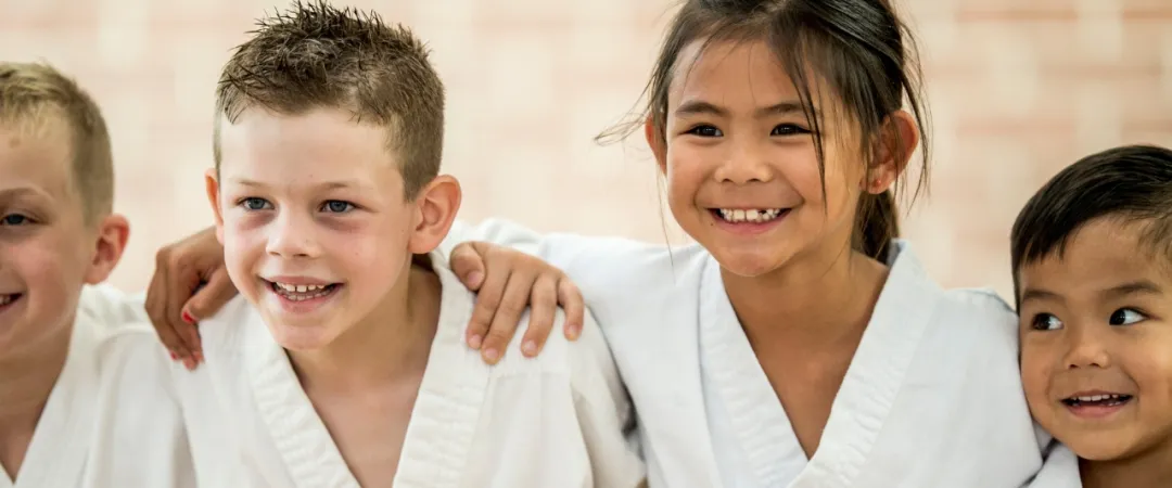 Four children in white karate uniforms stand with arms around each other, smiling brightly. They appear joyful and supportive in a martial arts setting.