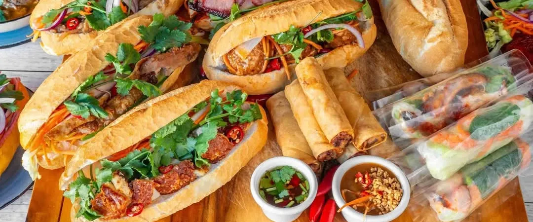 A wooden board filled with various Vietnamese banh mi sandwiches, crispy spring rolls, fresh rice paper rolls, dipping sauces, and a noodle bowl.