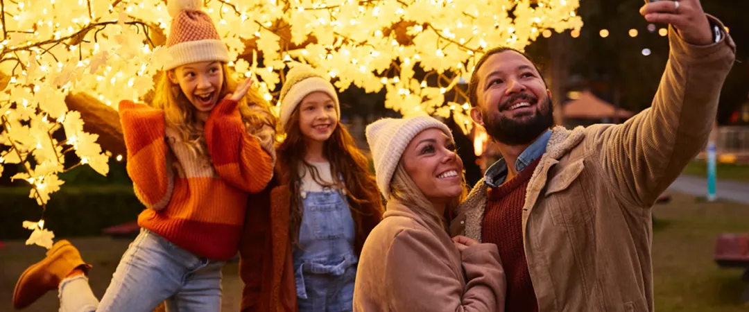 A joyful family taking a selfie under a glowing tree decorated with bright, star-shaped lights. They wear warm winter clothing, smiling and cheerful.