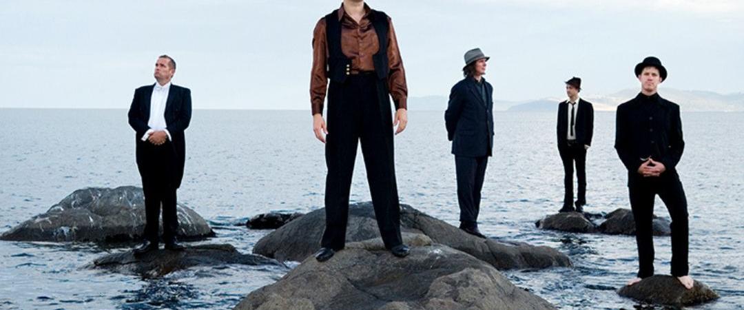 5 men in black suits standing on rocks in the ocean
