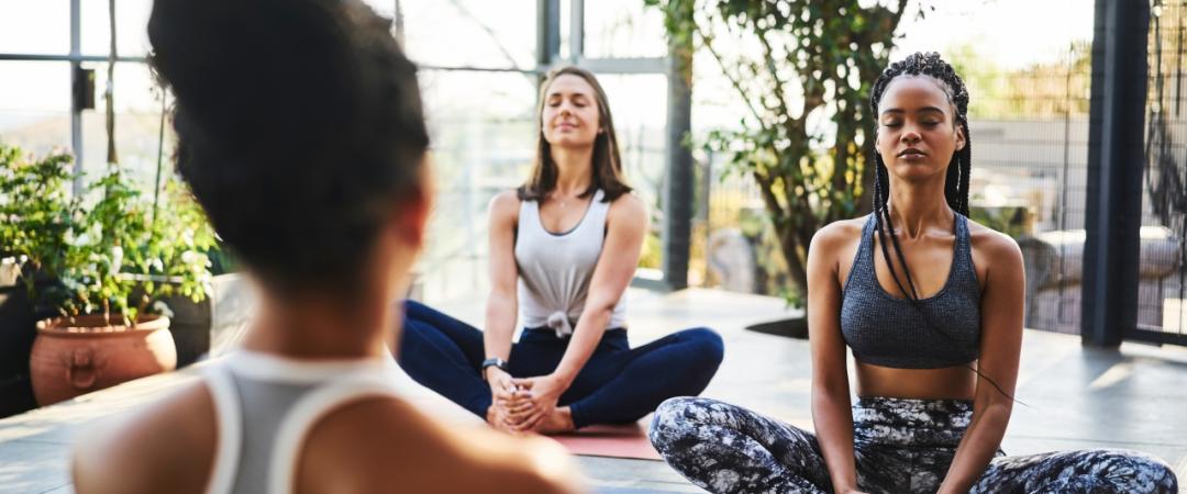 Three women practice yoga indoors, sitting cross-legged with eyes closed, appearing calm and focused. Sunlight filters through large windows and greenery surrounds them.