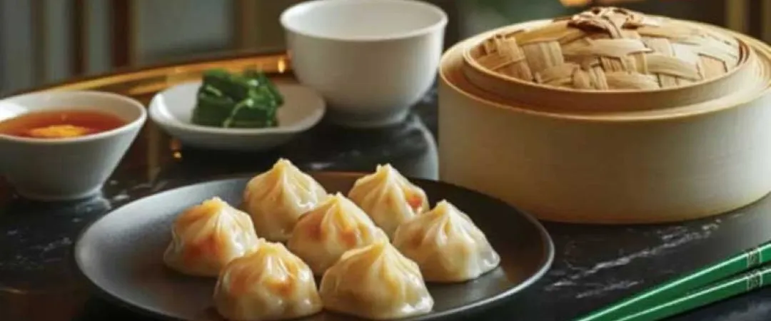 Dim sum spread with steamed soup dumplings, bamboo steamer basket, dipping sauce, tea, greens, and chopsticks in warm light.