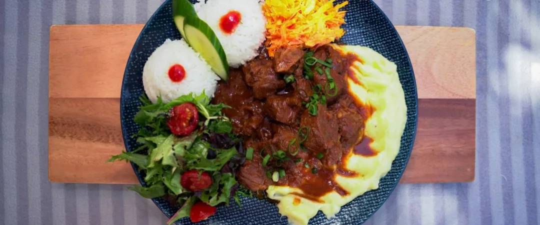 Overhead view of a colorful beef stew platter with mashed potatoes, rice, salad, tomatoes, and shredded carrots on a wooden board.