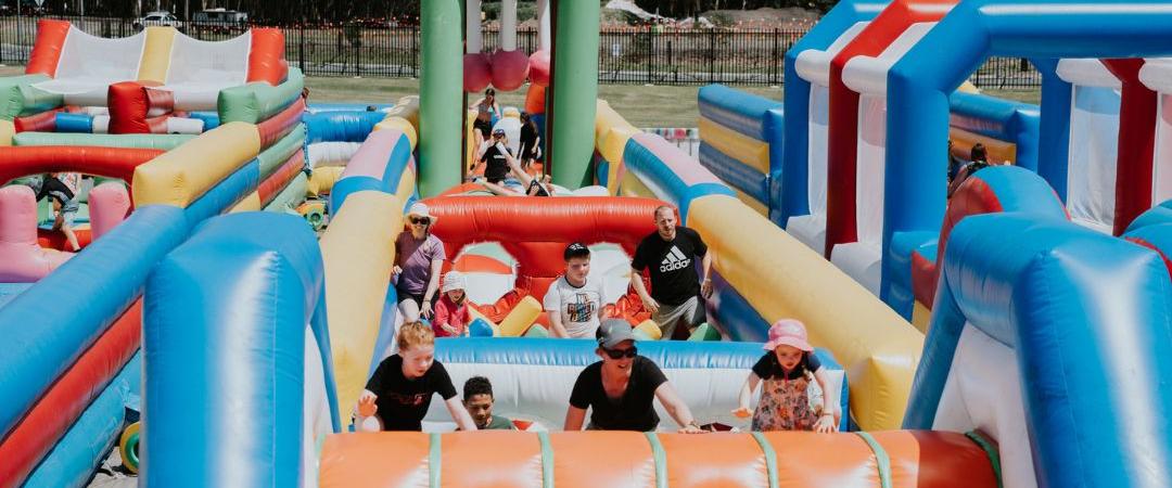A large, colorful inflatable obstacle course with slides and tunnels, surrounded by a crowd of people enjoying a sunny event. Energetic and vibrant atmosphere.