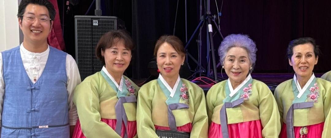 A group of five people pose together, four in colorful traditional Korean hanboks and one in a blue vest, smiling warmly in a festive setting.