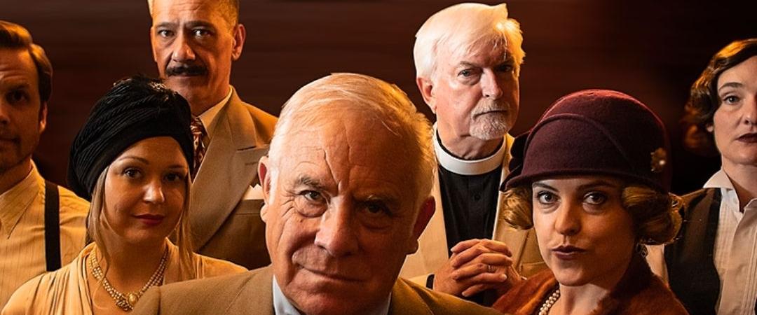 A group of seven diverse actors in period costumes, some with serious expressions, are posed together. The dim lighting gives a dramatic, vintage tone.
