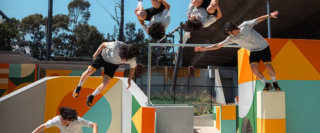 Parkour athletes leap and flip across colorful concrete obstacles in an outdoor urban course, captured midair against a sunny blue sky.