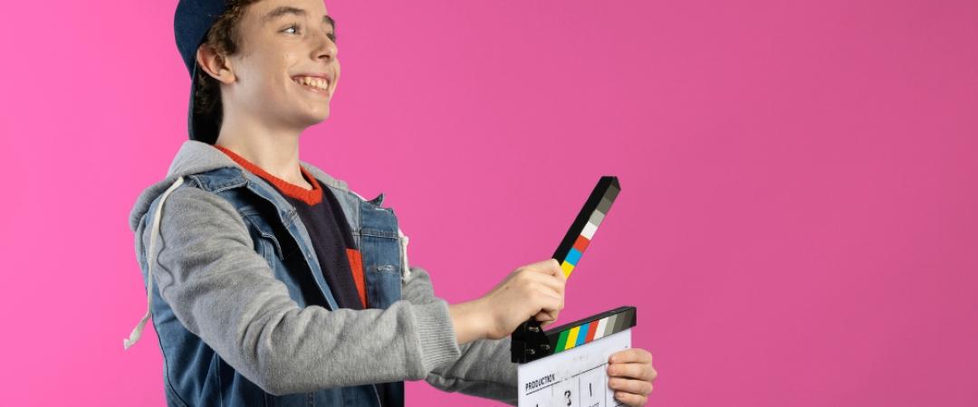 A smiling young person in a denim jacket and cap holds a clapperboard against a pink background. The mood is playful and creative.