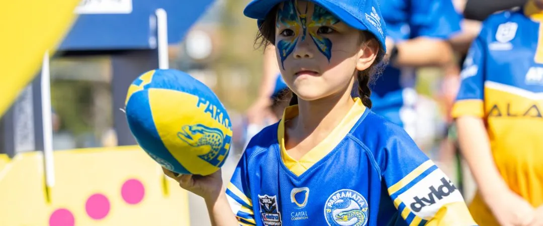 Little girl in blue and yellow hat & jersey throwing a ball. She's also got a blue butterfly on her face