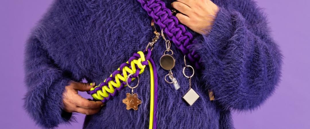 Person wearing a fuzzy purple sweater with chunky braided purple and yellow strap, adorned with various keychains and charms, against a purple background.