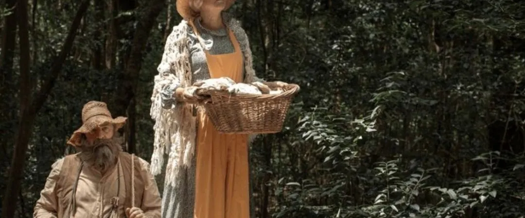 A woman in rustic attire stands, holding a basket near a bearded man with a drum, set in a forest. The scene conveys a folk, earthy ambiance.
