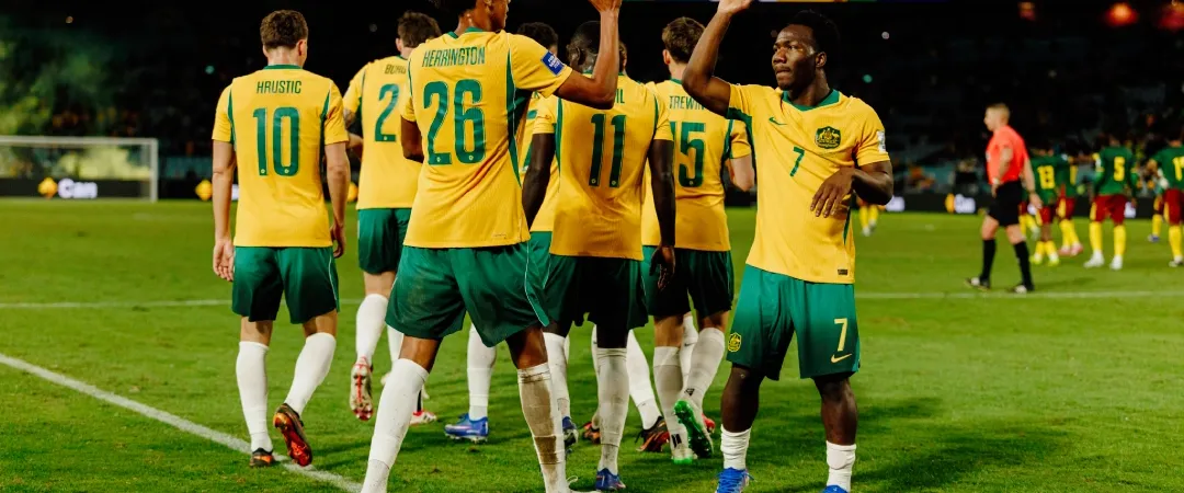 Soccer players in yellow and green uniforms high-five, celebrating during a night match. Other players and a scoreboard are visible in the background.