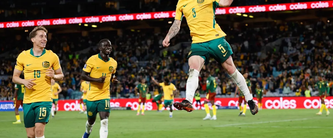 Soccer player in a yellow jersey leaps joyfully with arm raised, celebrating a goal. Teammates cheer, surrounded by a cheering crowd in a stadium.