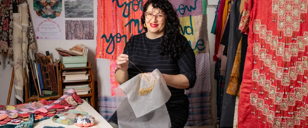 A woman sits in a colorful studio, holding embroidered fabric. She smiles, surrounded by textiles and a tapestry with bold lettering. Creative, warm atmosphere.