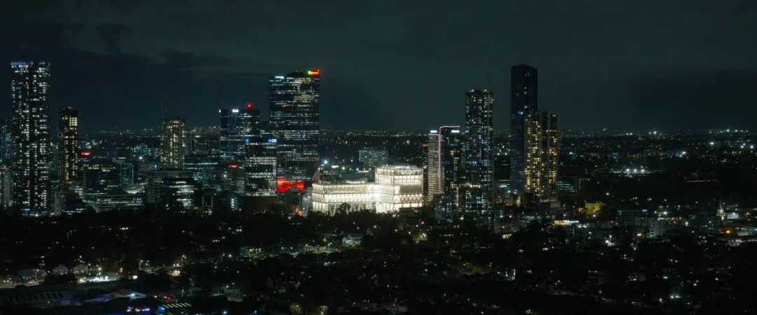 Nighttime city skyline with illuminated high-rise buildings and a bright white complex at center, viewed above dark neighborhoods beneath a cloudy sky.