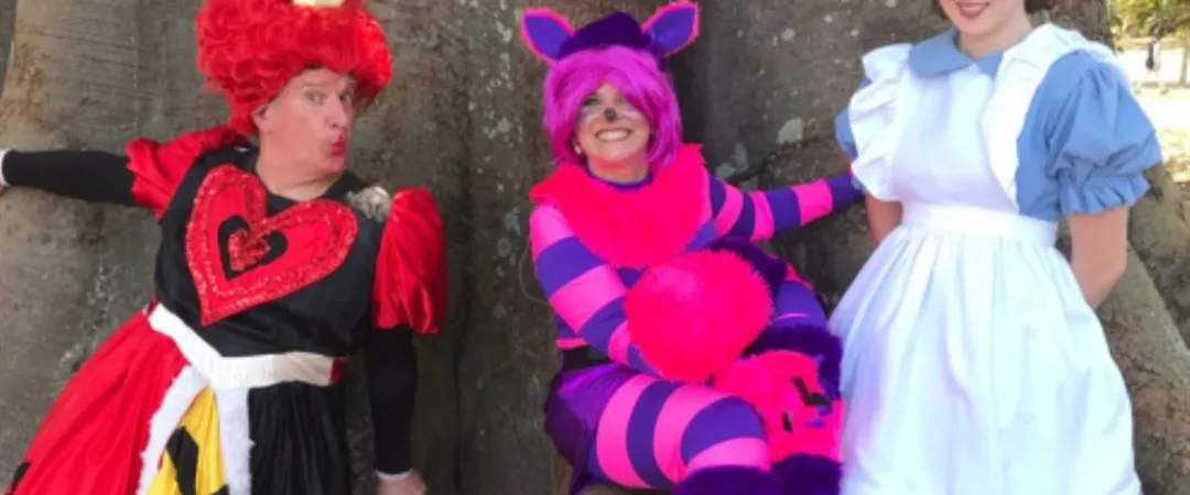 Three people in costume stand in front of a large tree. From left to right: the Queen of Hearts in red and black, the Cheshire Cat in pink and purple stripes, and Alice in a blue dress with a white apron. The scene is playful and whimsical, evoking a fantasy theme.