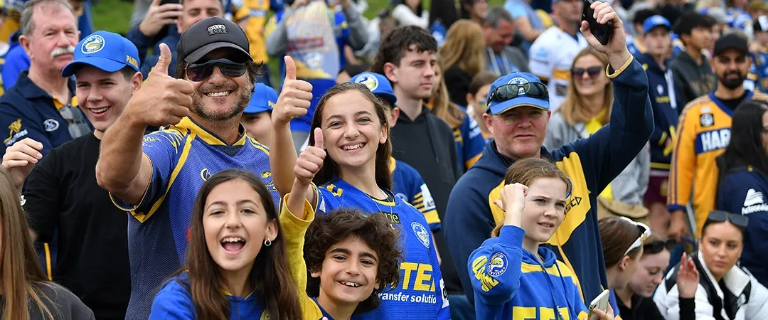 A joyful crowd wearing blue and yellow sports jerseys enthusiastically cheers and gives thumbs up, creating a lively and energetic atmosphere in a stadium.