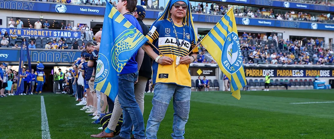 Fans stand on a soccer field, holding blue and yellow flags. The stadium is filled with cheering spectators, creating an energetic atmosphere.