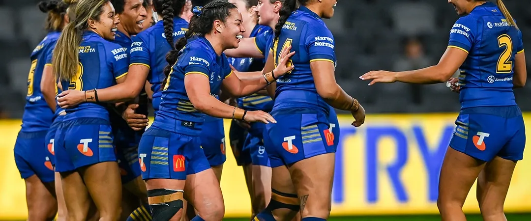 A women's sports team in blue jerseys celebrates jubilantly on a soccer field, hugging and high-fiving, conveying excitement and teamwork.