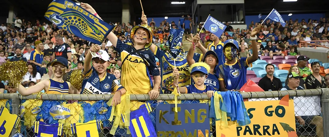 Crowd of enthusiastic football fans in blue and yellow jerseys, waving flags and cheering. A sign reads, "Go Parras! Let's Go." Vibrant, lively atmosphere.