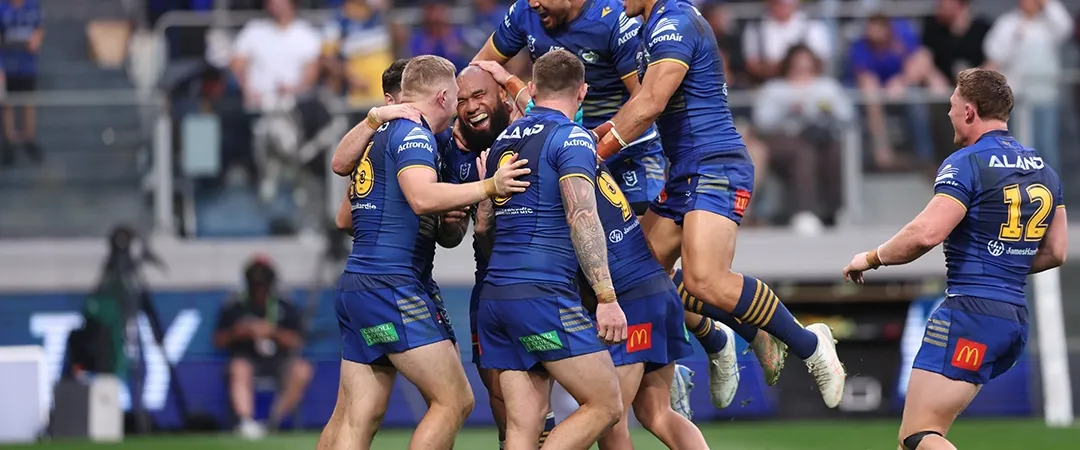 Rugby players in blue jerseys celebrate a try on the field, with one player lifted in the air. The crowd in the background is cheering enthusiastically.