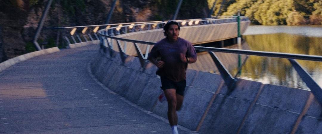 A person runs on a curving riverside path lined with railings, surrounded by lush trees. The scene conveys energy and tranquility in early morning light.