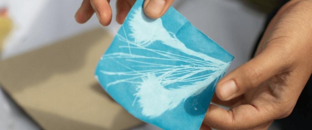 A person holds a blue, wrapped sanitary pad above a table during daylight. Nearby are a cardboard box and some scattered objects, suggesting an educational or casual setting.
