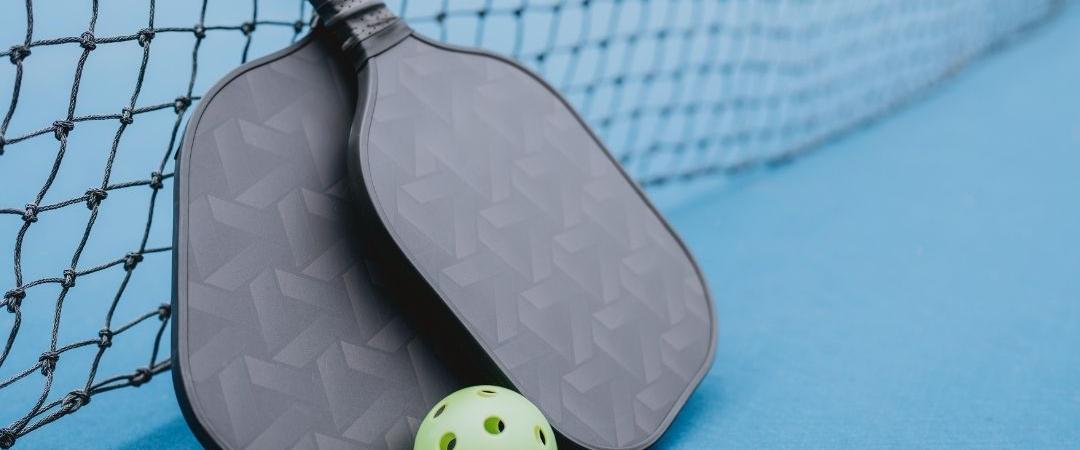 Two pickleball paddles and a green pickleball rest against a black net on a blue court. The scene conveys readiness and anticipation for play.