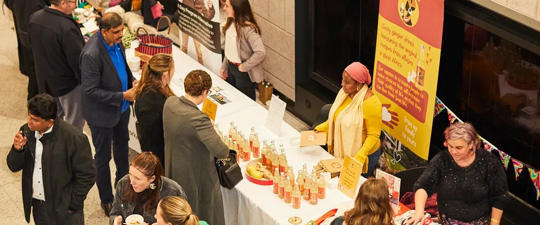 People browse colourful stalls displaying bottled drinks, crafts and other products at an indoor community marketplace.