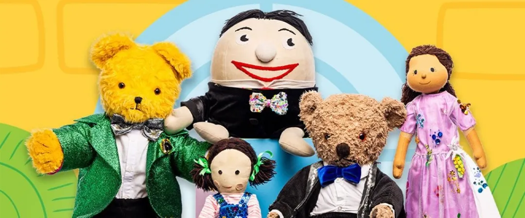 Five colorful dolls are seated against a playful yellow and blue backdrop. The scene is cheerful, with each doll dressed in fancy outfits.