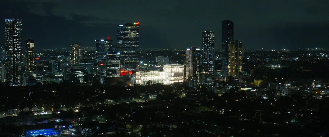 A sprawling city skyline glows at night, with illuminated high-rises and a bright central building beneath a dark, cloudy sky, creating a moody urban scene.