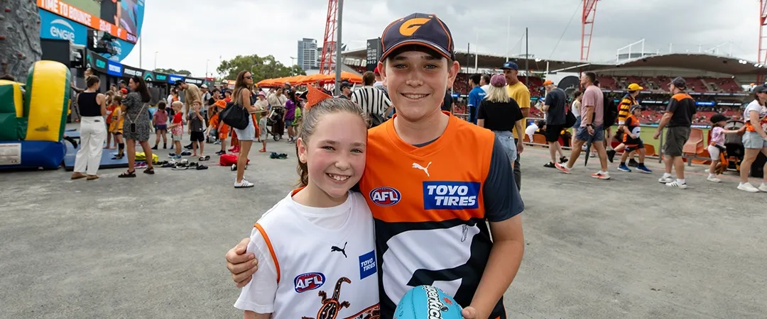 Two smiling kids at an outdoor event, wearing AFL-themed shirts. One holds a football. Crowds and a sports stadium are visible in the background.