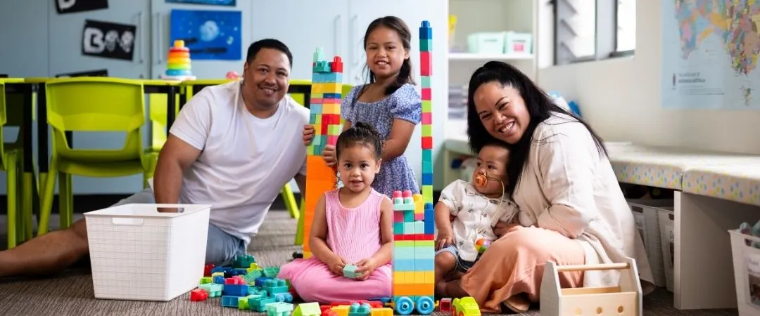 A smiling family of five sits on the floor of a colourful playroom, surrounded by toys and building blocks.