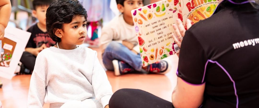 A young child attentively listens to an adult reading a colorful book titled "I Can Eat a Rainbow" in a classroom setting with other kids sitting nearby.