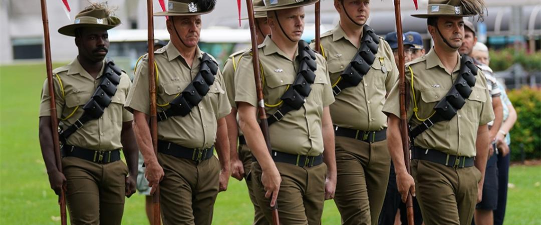 A group of uniformed soldiers marches in formation on grass, holding flags. They wear hats and sashes. The setting is formal, conveying discipline.