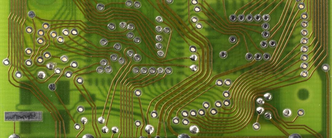 Close-up image of a green circuit board with intricate copper traces and scattered solder points, conveying complexity and technological precision.