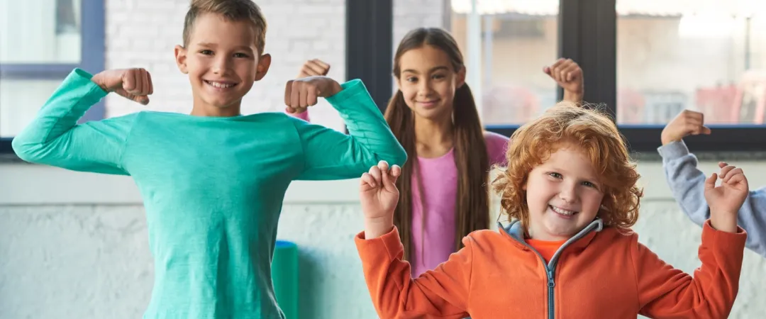 Four smiling children in colorful clothes strike fun muscle poses indoors, conveying joy and confidence. Bright window light in the background.